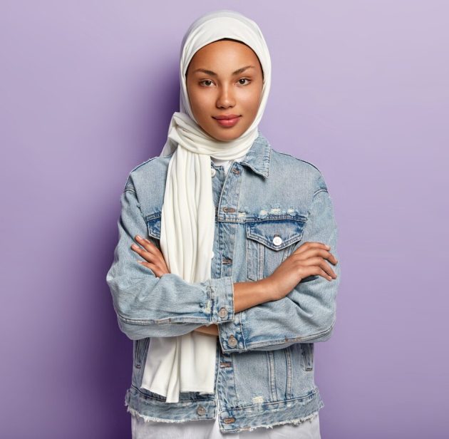 attractive-eastern-woman-covers-head-with-white-headscarf-guard-her-dignity-power-has-special-dress-code-keeps-hands-crossed-looks-with-modesty-poses-purple-wall-islamic-rules_273609-26323